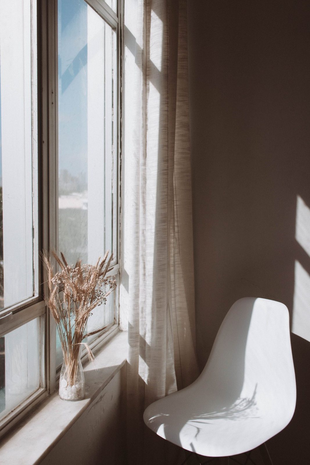 Ein leerer weißer Stuhl neben einem Fenster mit Vorhang, davor eine Vase mit Trockengräsern im Sonnenlicht