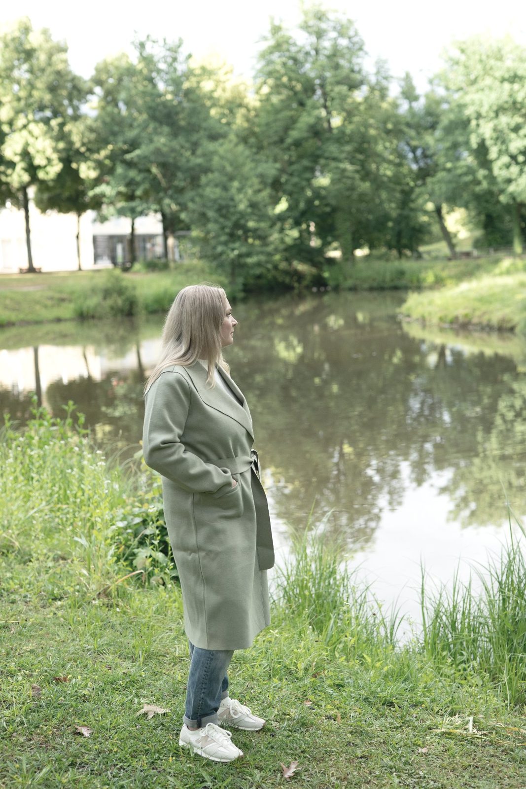 Mandy Jäger steht in einem salbeigrünen Mantel am Ufer eines Teiches und blickt über das Wasser