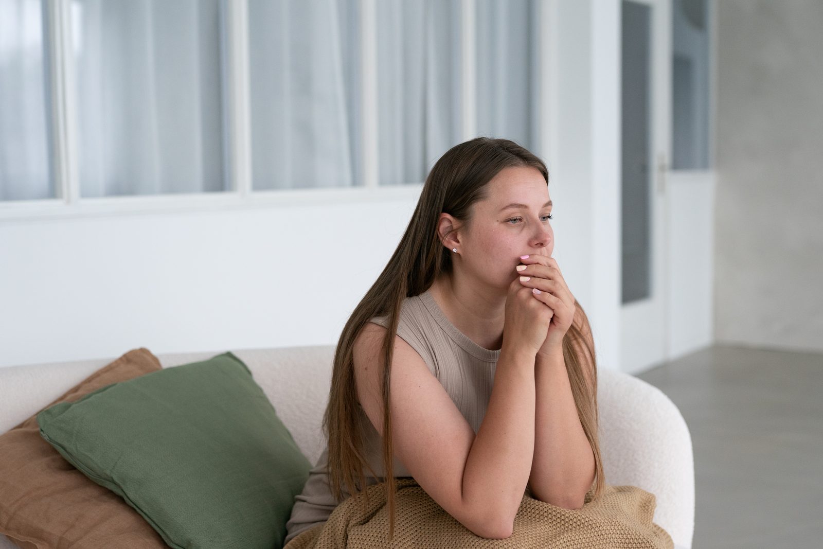 Eine Frau sitzt nachdenklich auf einem hellen Sofa und schaut zur Seite